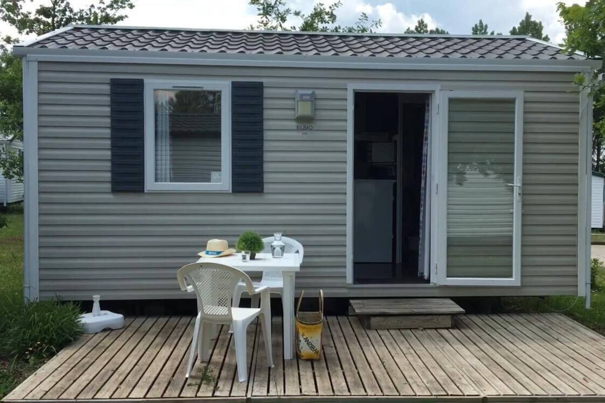 Una casa móvil de una sola planta, pequeña, con una puerta corrediza, un patio pequeño con mesa y sillas, y una terraza de madera.