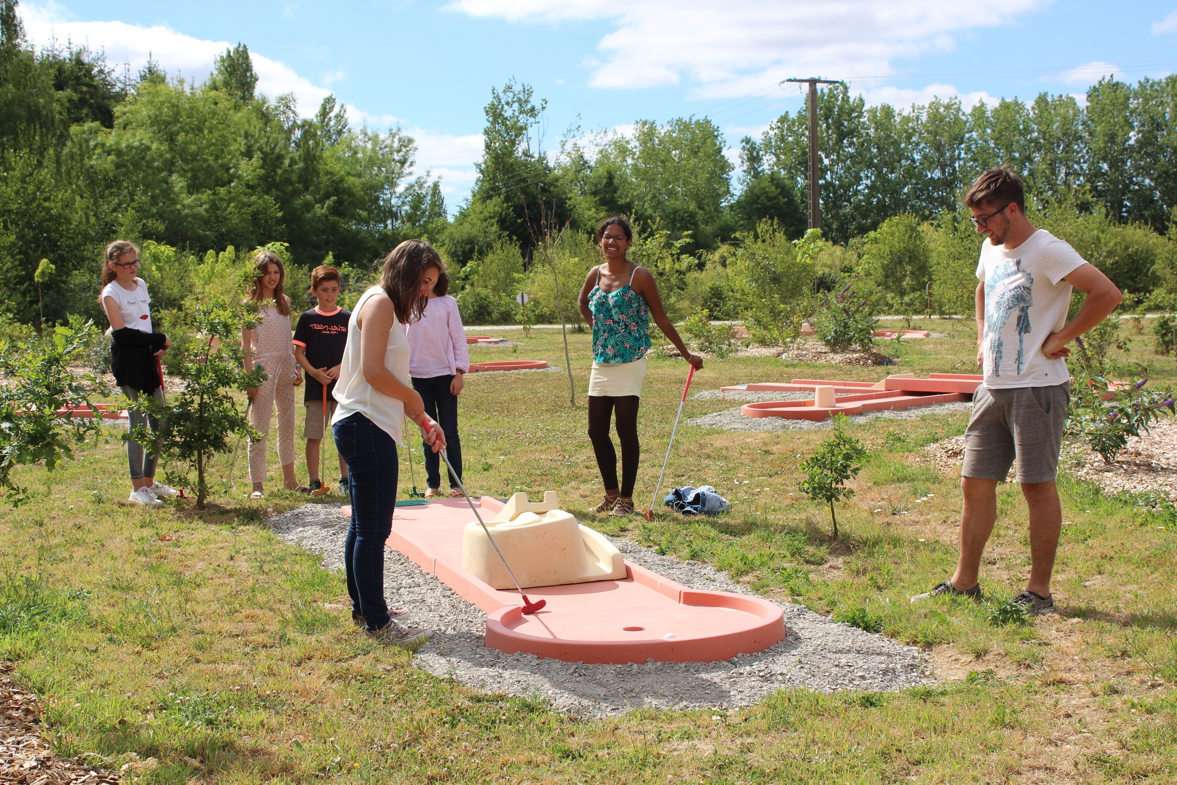 A group of people playing mini-golf in an outdoor setting with a red and white golf course, surrounded by greenery and trees.