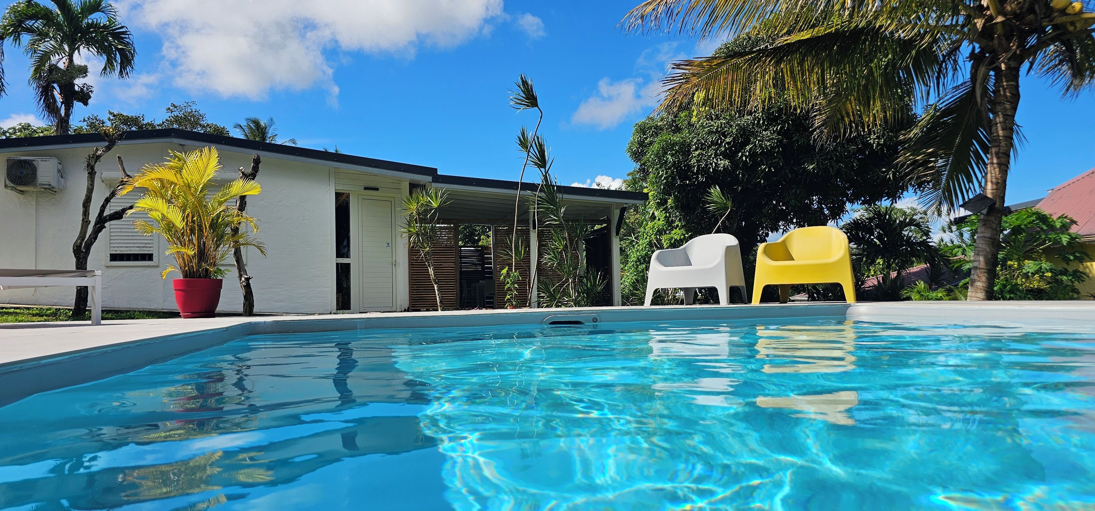 A swimming pool with clear blue water, surrounded by a white building with a covered patio area, and two plastic chairs.