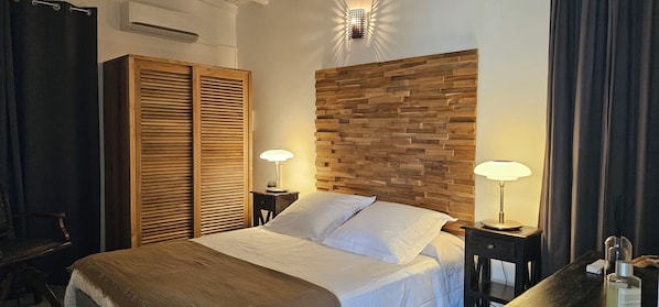 A hotel room with a bed, wooden headboard, two bedside lamps, a wardrobe, and a small table with a lamp and a bottle. - Habitation Villa Les Cassias (Petit-Bourg)