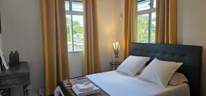 A neatly made bed with white linens and pillows, a bedside table with a lamp, and a window with curtains. - Habitation Villa Les Cassias (Petit-Bourg)