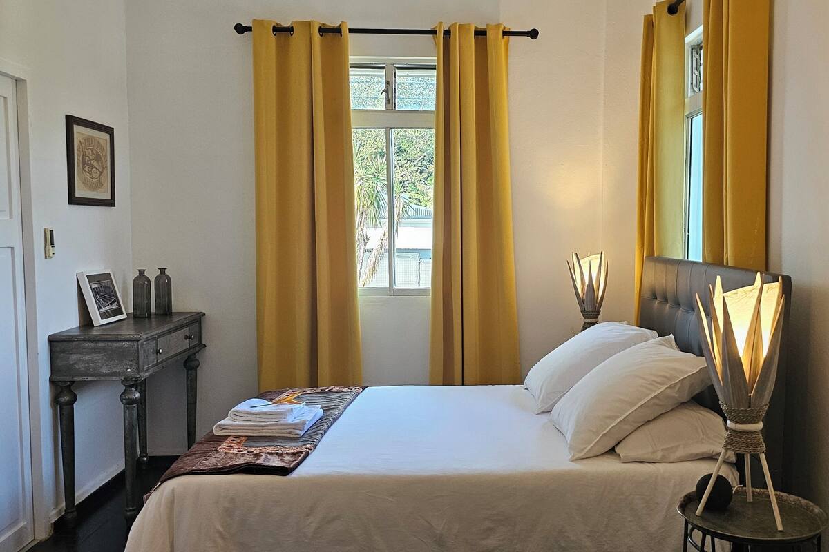 A hotel room with a bed, a bedside table with a lamp, a window with curtains, and a framed picture on the wall.