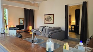 Un saló modern amb un sofà gris, una taula de centre de fusta, una zona de cuina amb un rentatge i un mirall gran a la paret.