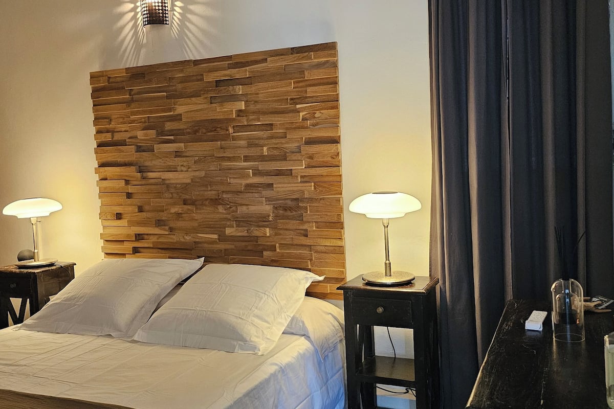 A neatly made bed with white linens, two bedside lamps, a wooden headboard, and a mirror with a decorative item on the wall.