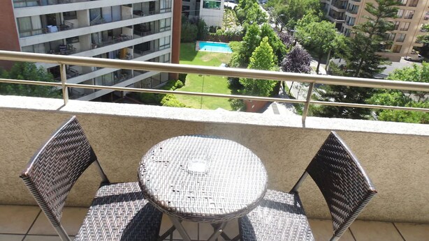 Un balcón con una mesa redonda y dos sillas de mimbre, con vista a una zona residencial con varios edificios de apartamentos y un espacio verde con piscina.