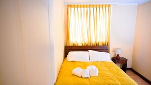 Habitación de hotel con colcha amarilla, almohadas blancas y cabecero de madera. Se observa un mesita de noche con una lámpara y un rollo de papel higiénico.