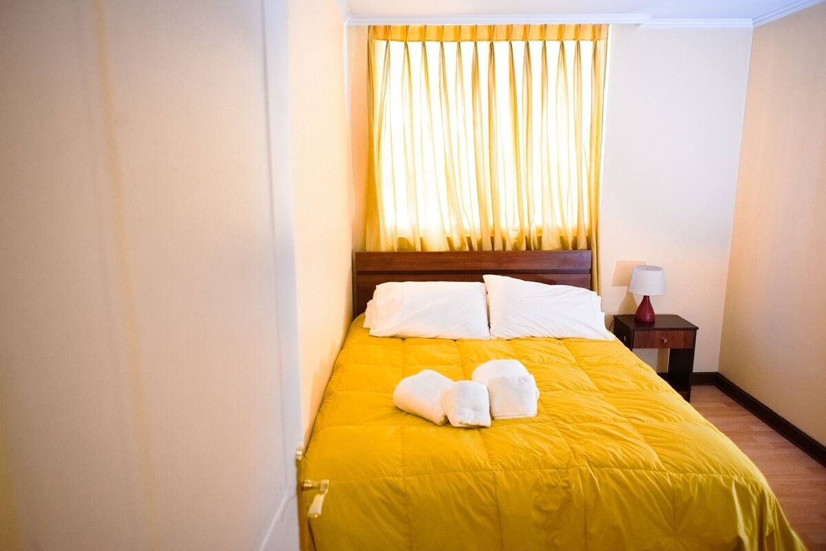 Ein Hotelzimmer mit gelber Bettdecke, weißen Kissen und einem Holz-Kopfteil. Es gibt einen Nachttisch mit einer Lampe und einer Toilettenpapierrolle.