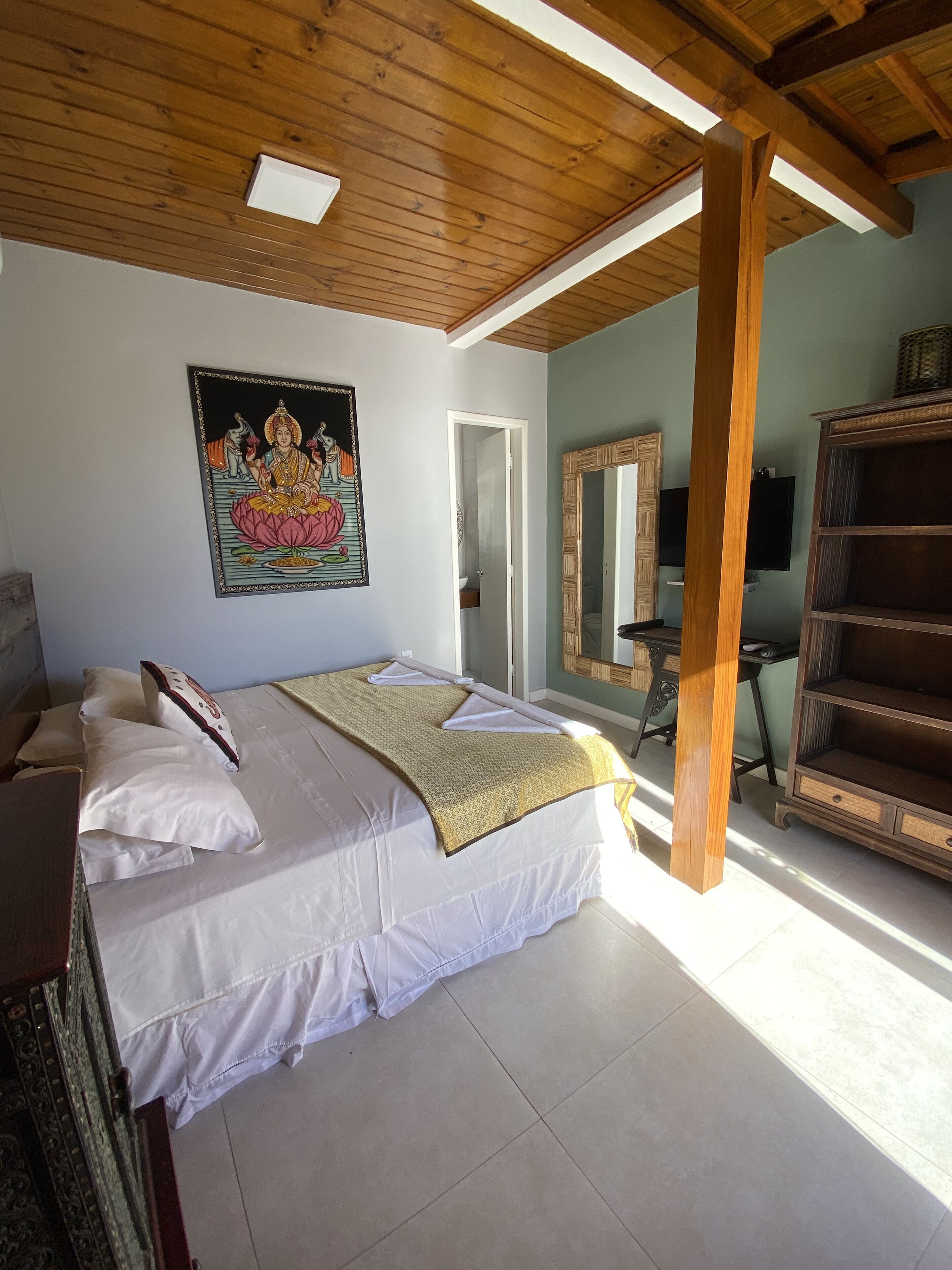 A bedroom with a large bed, a wooden ceiling, a painting on the wall, a mirror, and a TV.