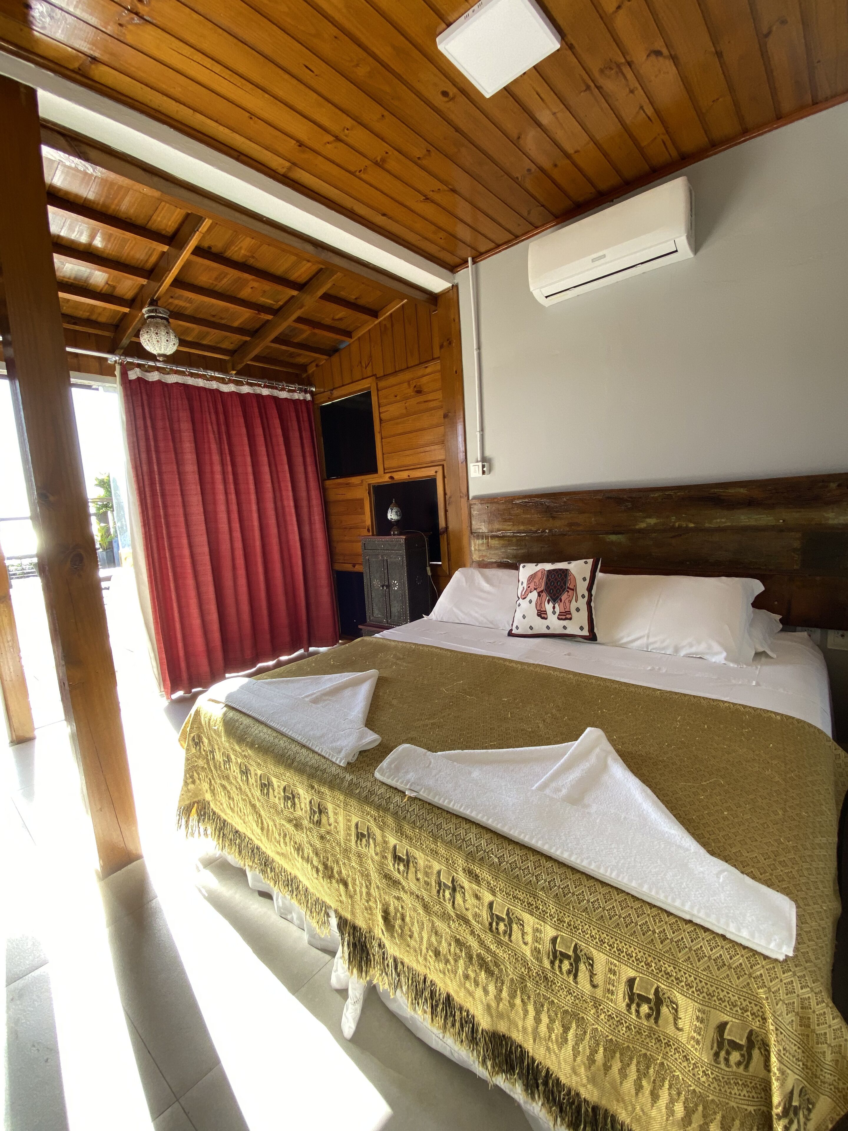 Une chambre d’hôtel avec un plafond en bois, un lit recouvert d’une couverture à motifs, des oreillers blancs et un rideau rouge.