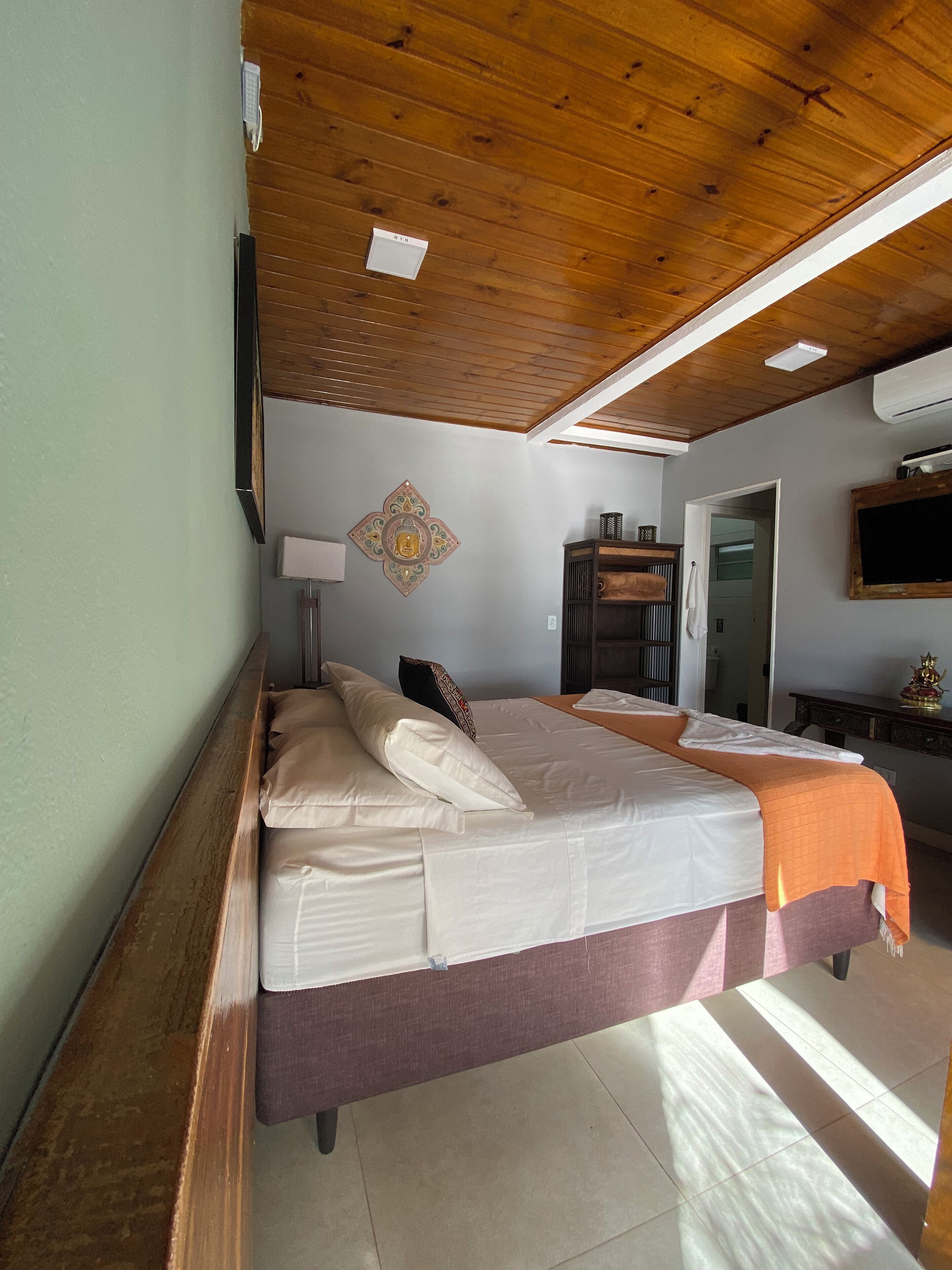 A bedroom with a wooden ceiling, a bed with white and orange bedding, a wall-mounted TV, a decorative wall piece, and a desk with a mirror and a small statue.