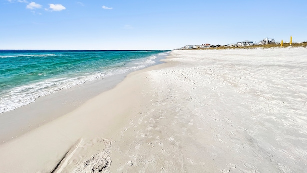 A beach with clear blue water, sandy shore, and houses in the background.