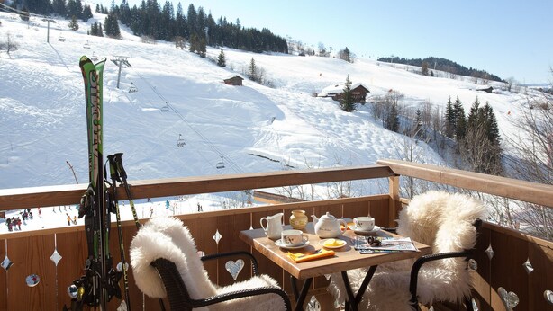 Une terrasse en bois avec une table préparée pour le thé, des paires de skis appuyés contre le mur et une vue imprenable sur une montagne enneigée.