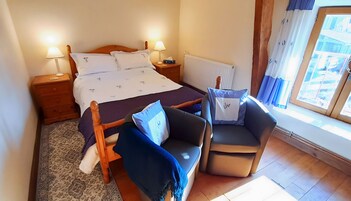Un dormitorio con cama, dos mesitas de noche, una mesita auxiliar, una silla, una ventana con cortinas y un radiador.
