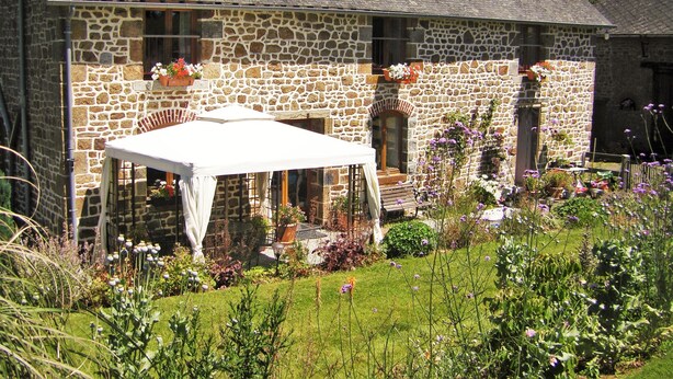 A stone house with a covered patio, a garden with various plants and flowers, and a clear sky.