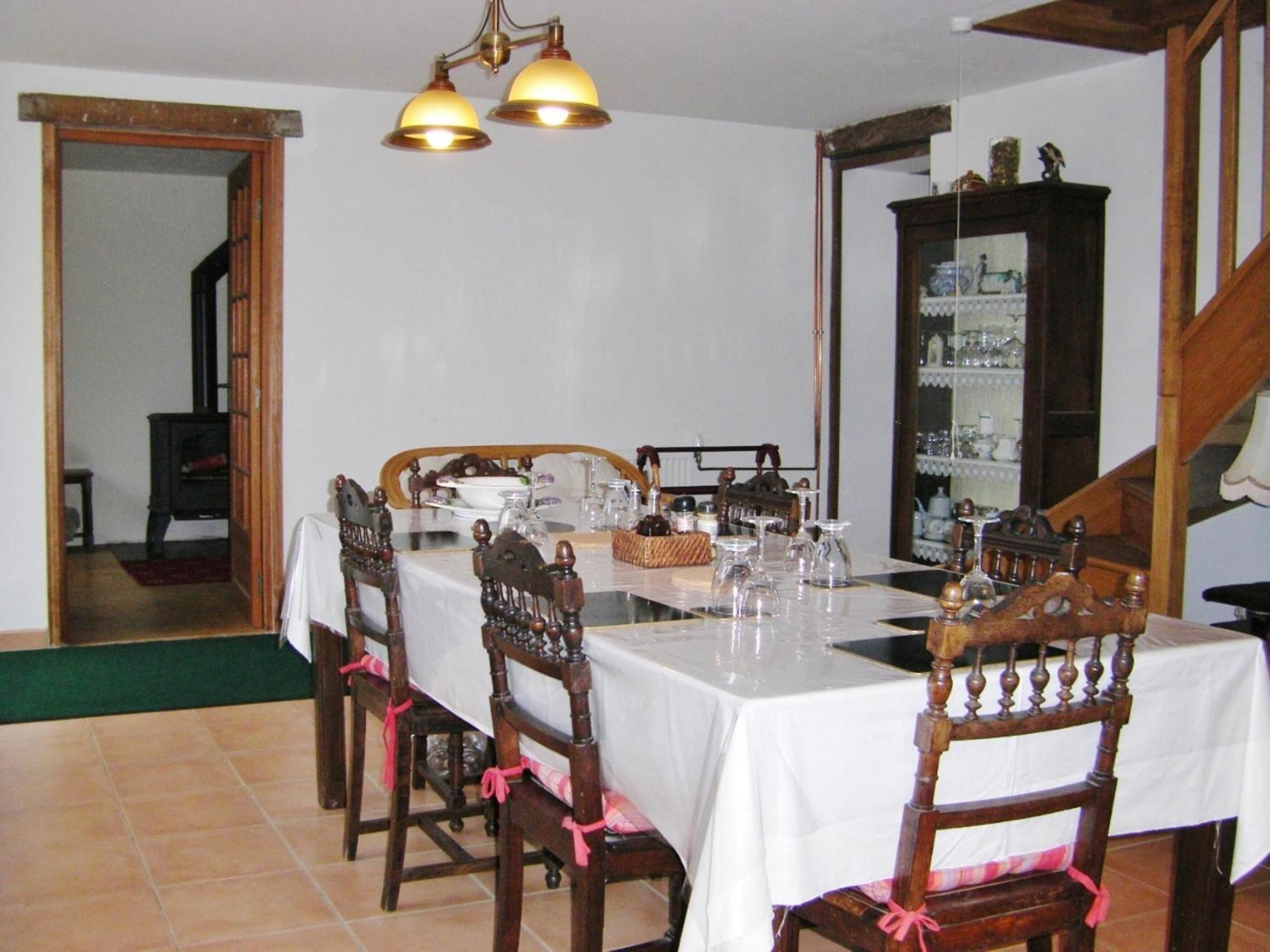Une salle à manger avec une longue table dressée pour six personnes, des chaises en bois ornées de rubans roses, un lustre et un buffet en bois contenant de la verrerie.