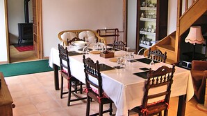 A dining area with a table set for four, surrounded by wooden chairs. A chandelier hangs above, and a bookshelf filled with books and decorative items is visible in the background.
