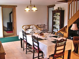 Un comedor con una mesa dispuesta para cuatro personas, rodeada de sillas de madera. Un candelabro cuelga encima, y se distingue una estantería llena de libros y objetos decorativos al fondo.