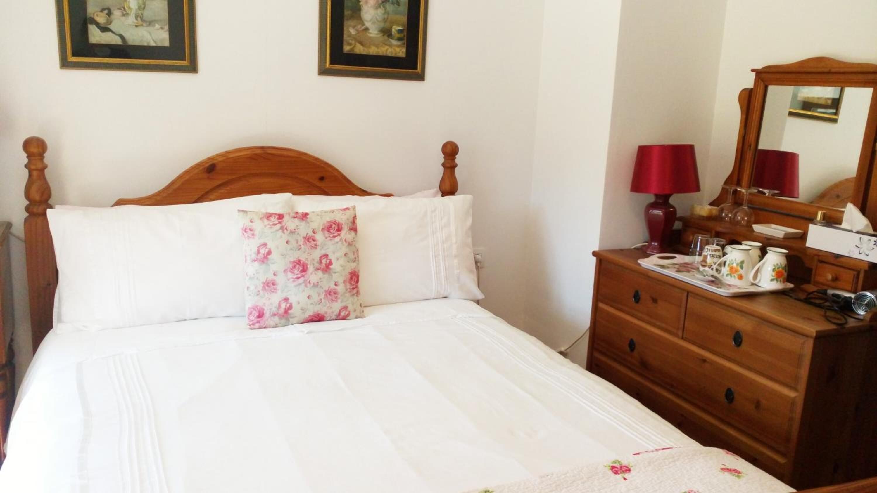 A neatly made bed with a floral pillow, a wooden dresser with a mirror, and a red lamp.