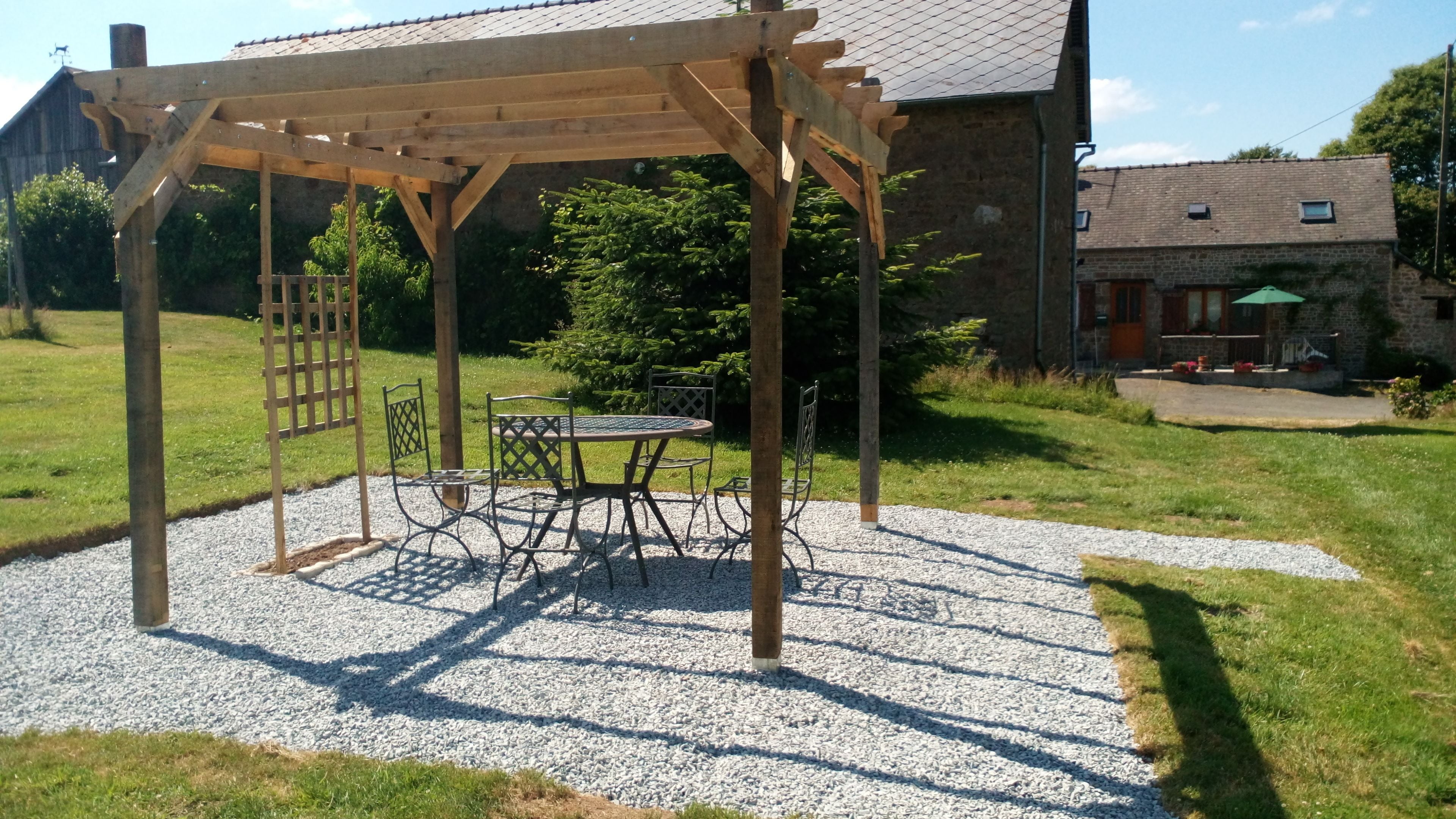 A wooden pergola with a table and chairs in a garden area with gravel flooring.