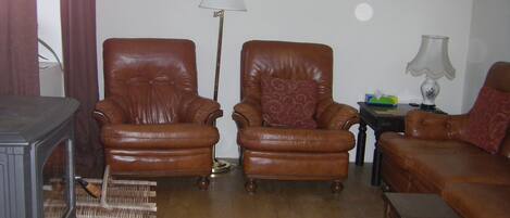 Una sala de estar con dos sillones de cuero marrón, una mesa de centro de madera, un tapete rojo estampado, una lámpara de pie y una mesita con lámpara.