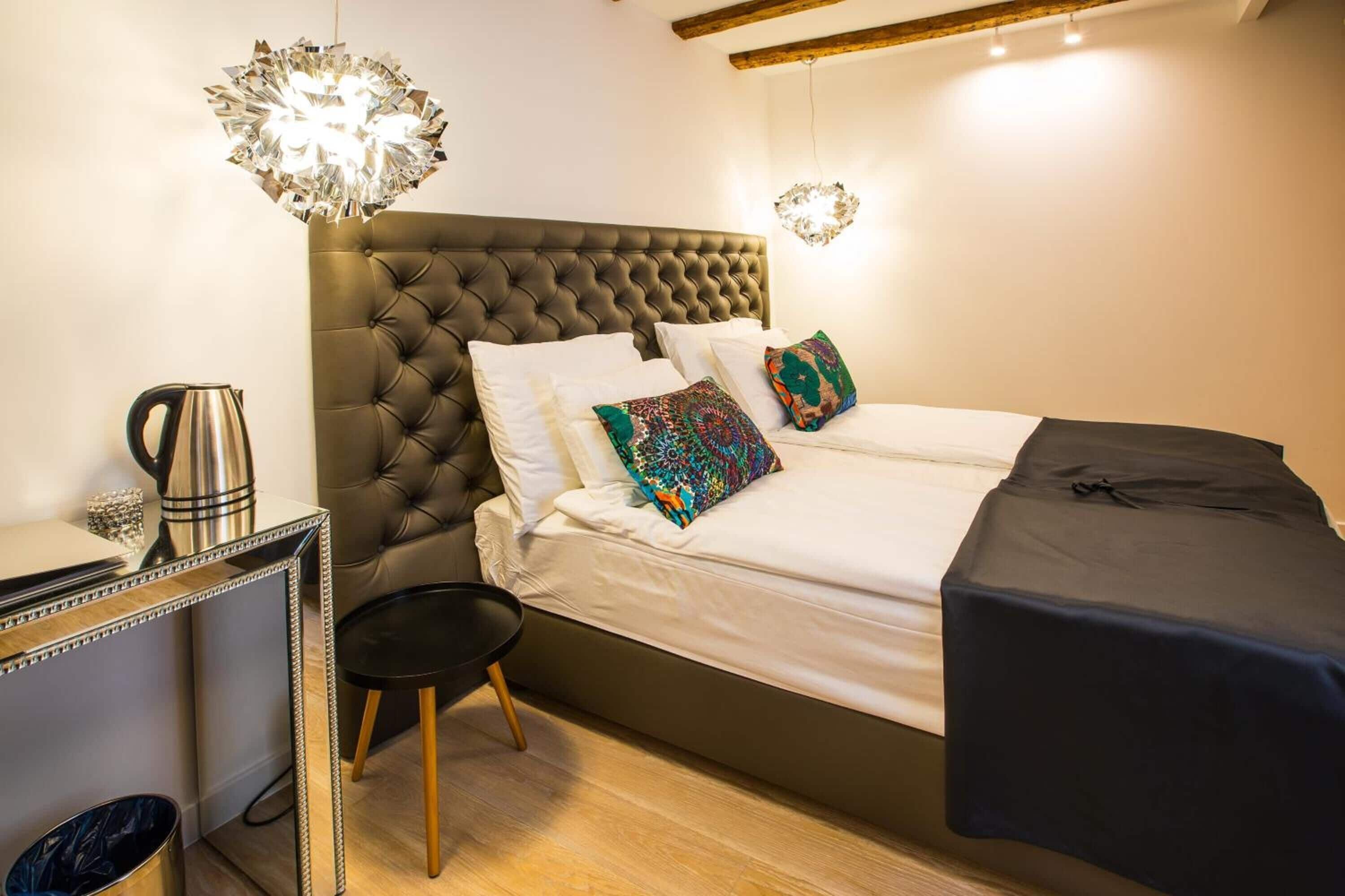 A bedroom with a tufted headboard, a bed with white linens and colorful pillows, a small black side table, a kettle on a side table, and a mirror with a decorative frame.