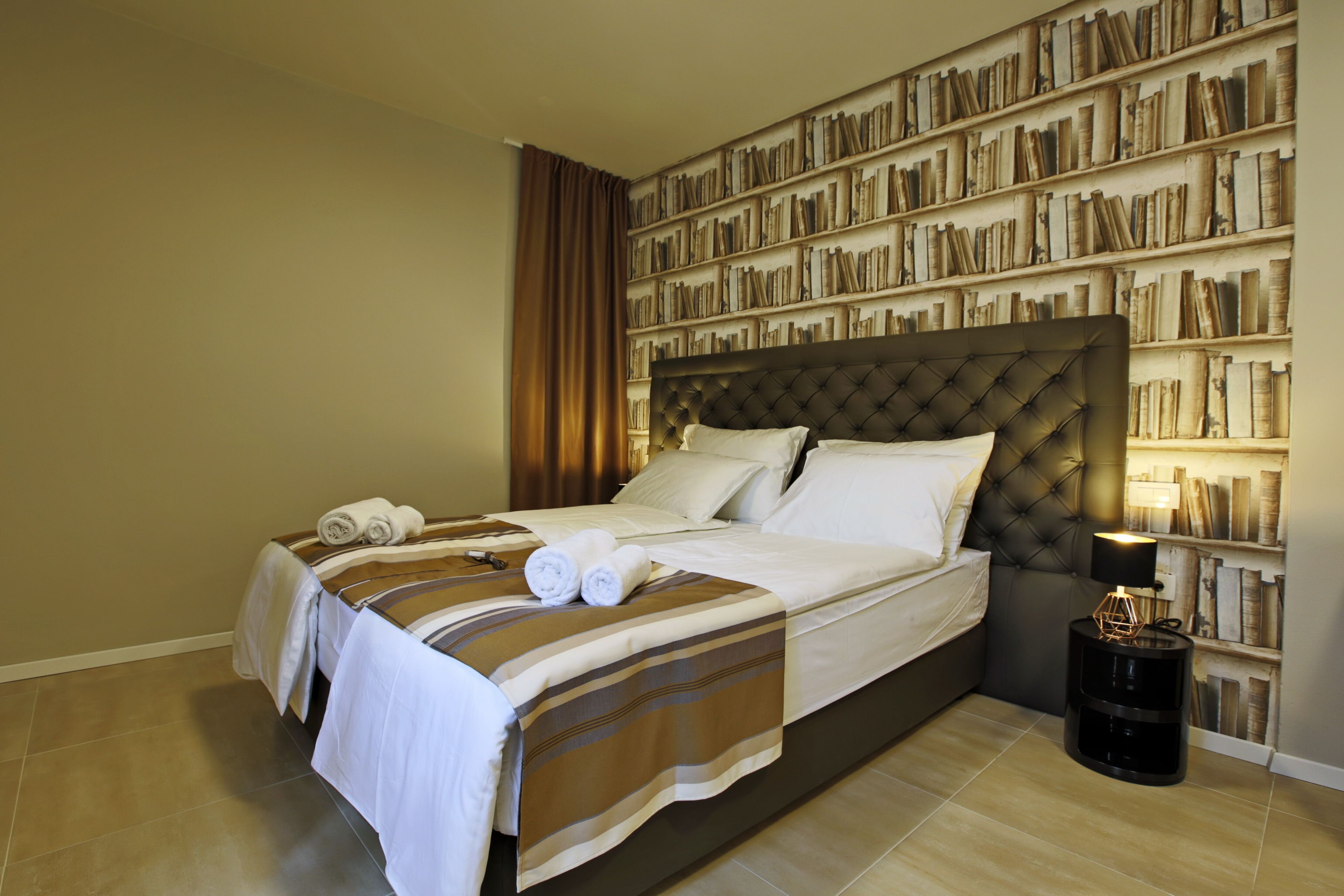 A neatly made bed with a striped bedspread, a bedside table with a lamp, and a bookshelf filled with books.