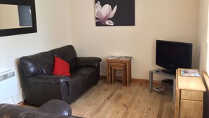 A living room with a black leather sofa, a small wooden table with magazines, a TV on a stand, and a framed picture of a flower on the wall. - One bedroom Wyvis (Inverness)