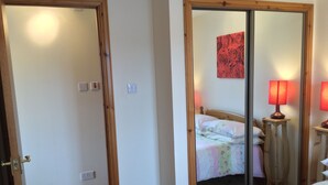 A bedroom with a wooden door, a bed with floral bedding, a nightstand with a lamp, and a mirror reflecting the room. - One bedroom Wyvis (Inverness)