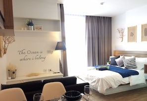 A hotel room with a bed, a desk with a lamp, a dining area with chairs and a table, and a wall with a decorative quote. - Simply Comfy Apartment by Patsamon (Pattaya)