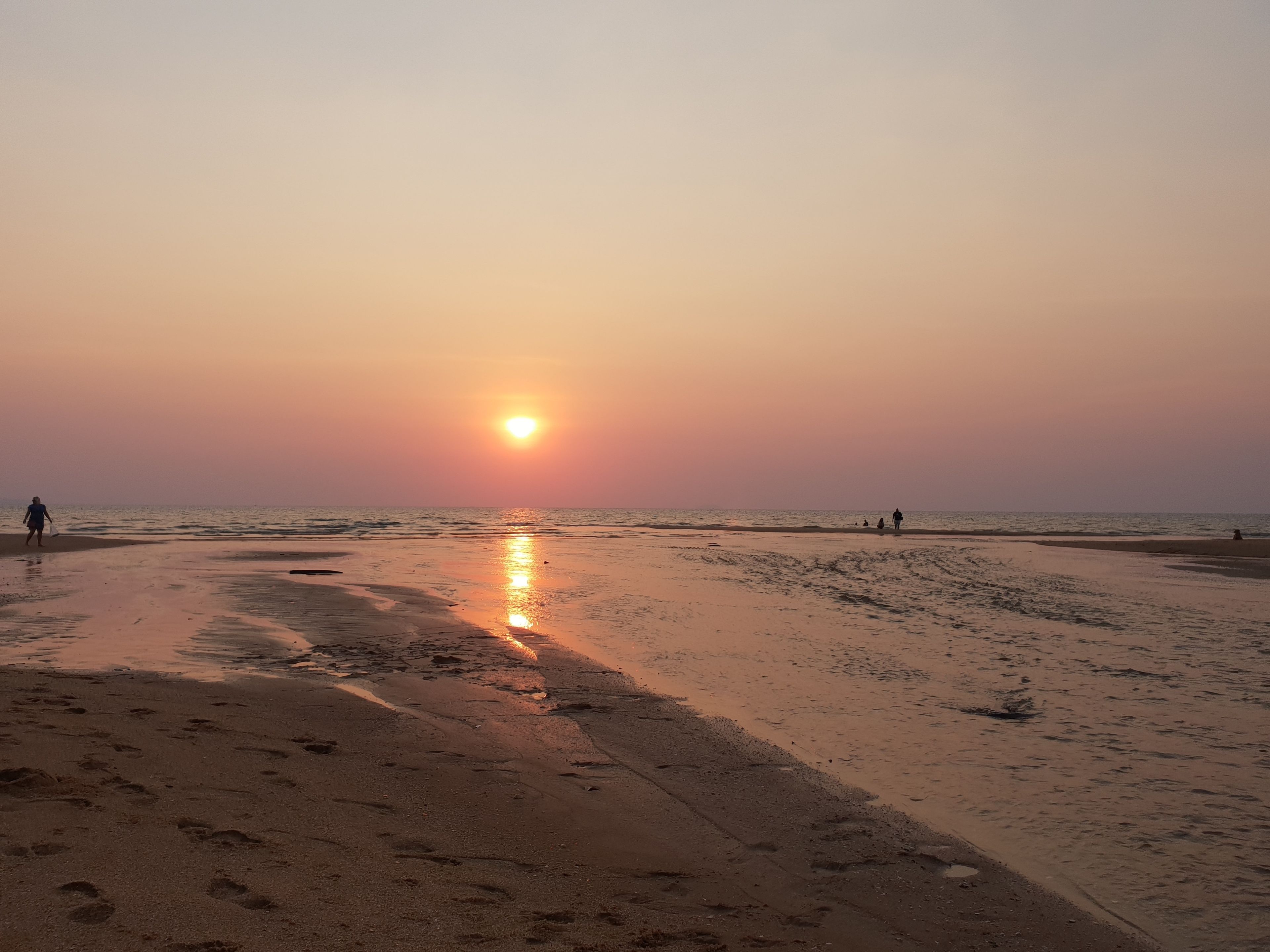 A beach at sunset with footprints in the sand and a few people walking.