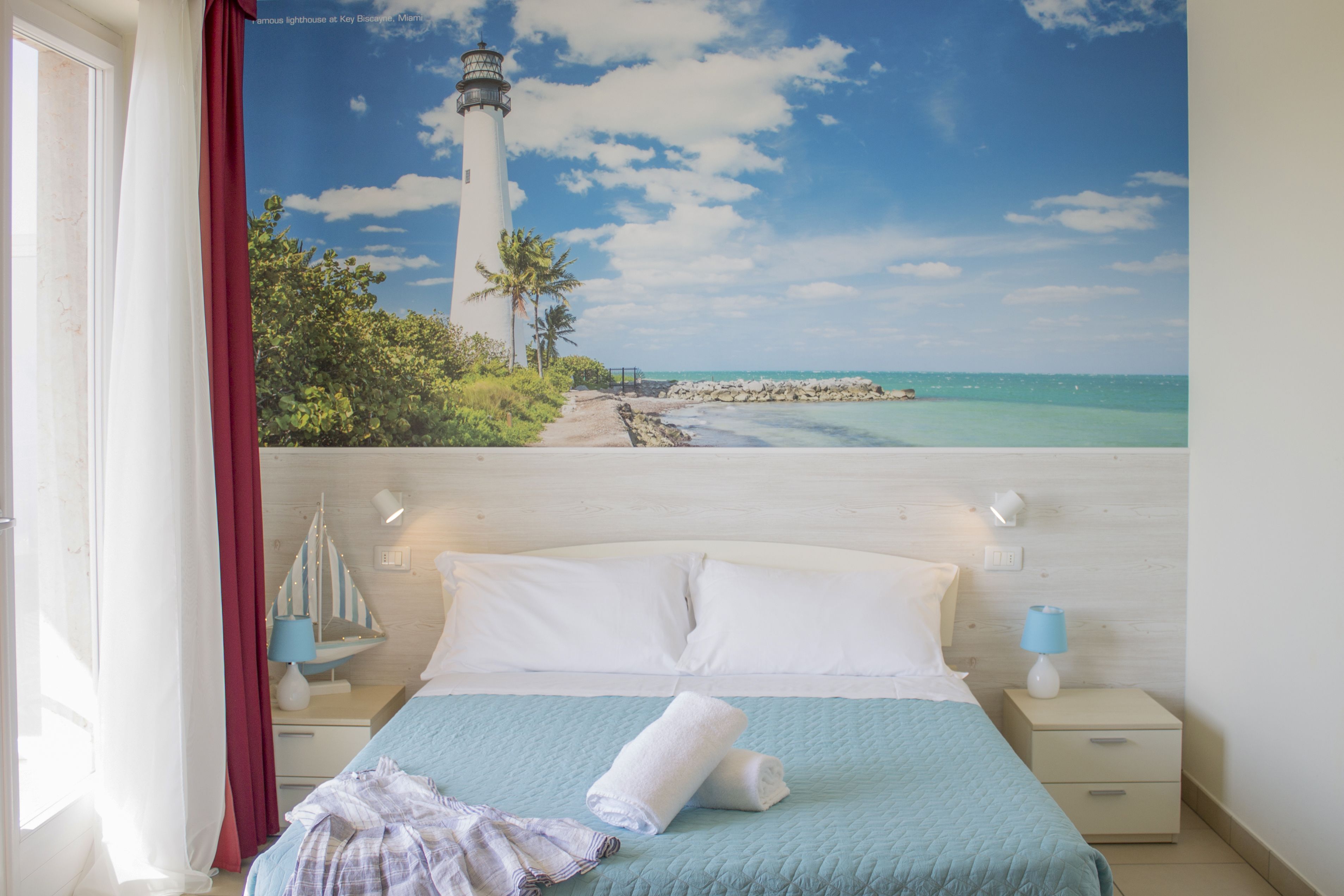 A bedroom with a bed, a nightstand, a lamp, and a view of a lighthouse and beach.
