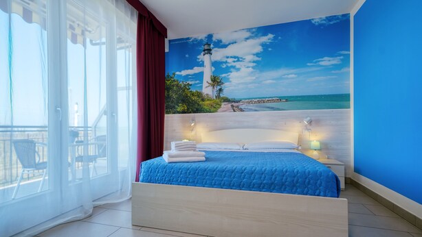 Ein Schlafzimmer mit Bett, Nachttisch, Lampe, einem Fenster mit Vorhängen und Blick auf einen Strand mit Leuchtturm.