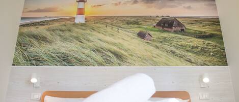 A bedroom with a bed, a wall-mounted light, and a painting of a lighthouse and landscape.