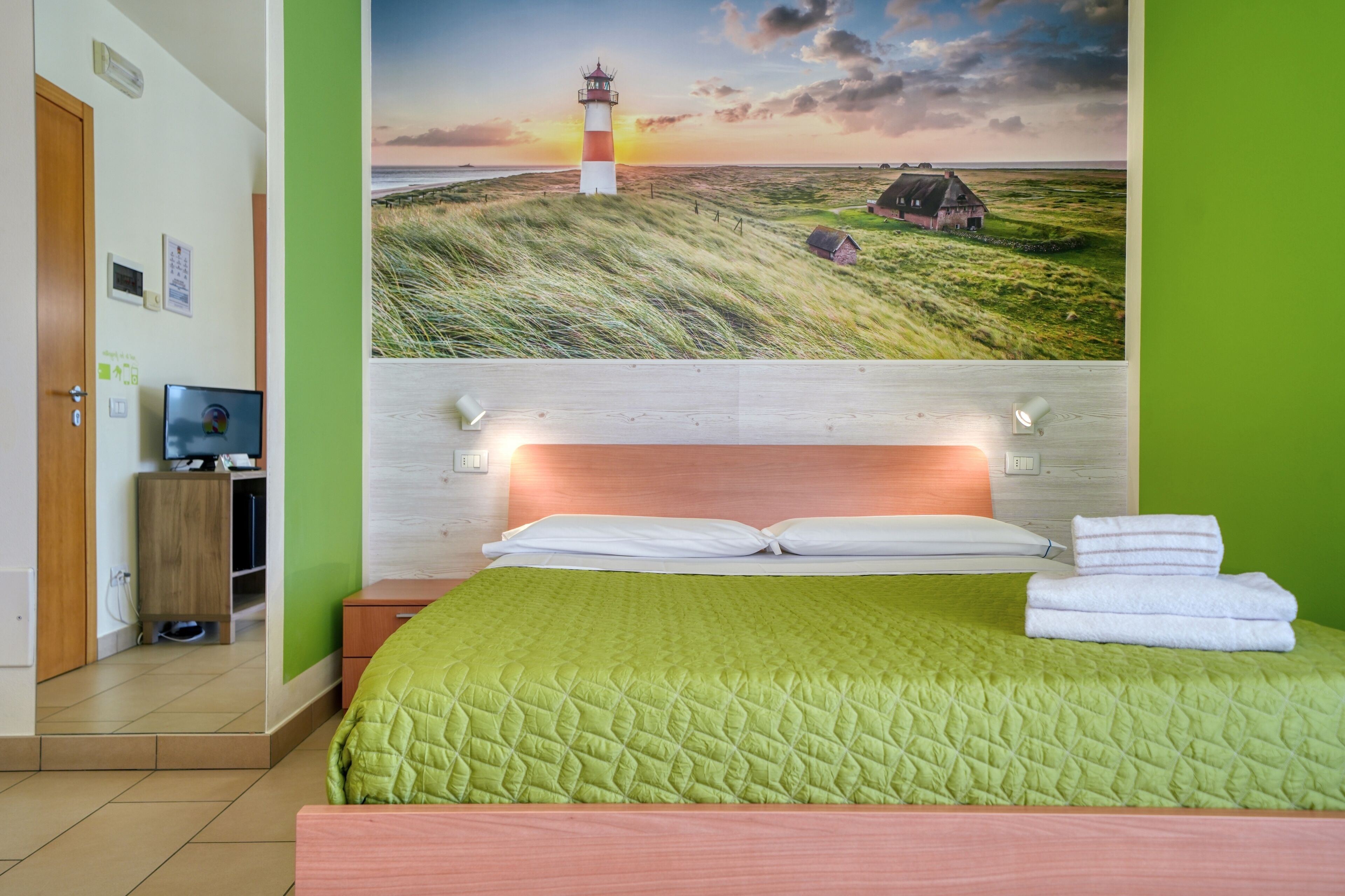 A hotel room with a bed, a TV, and a painting of a lighthouse on the wall.