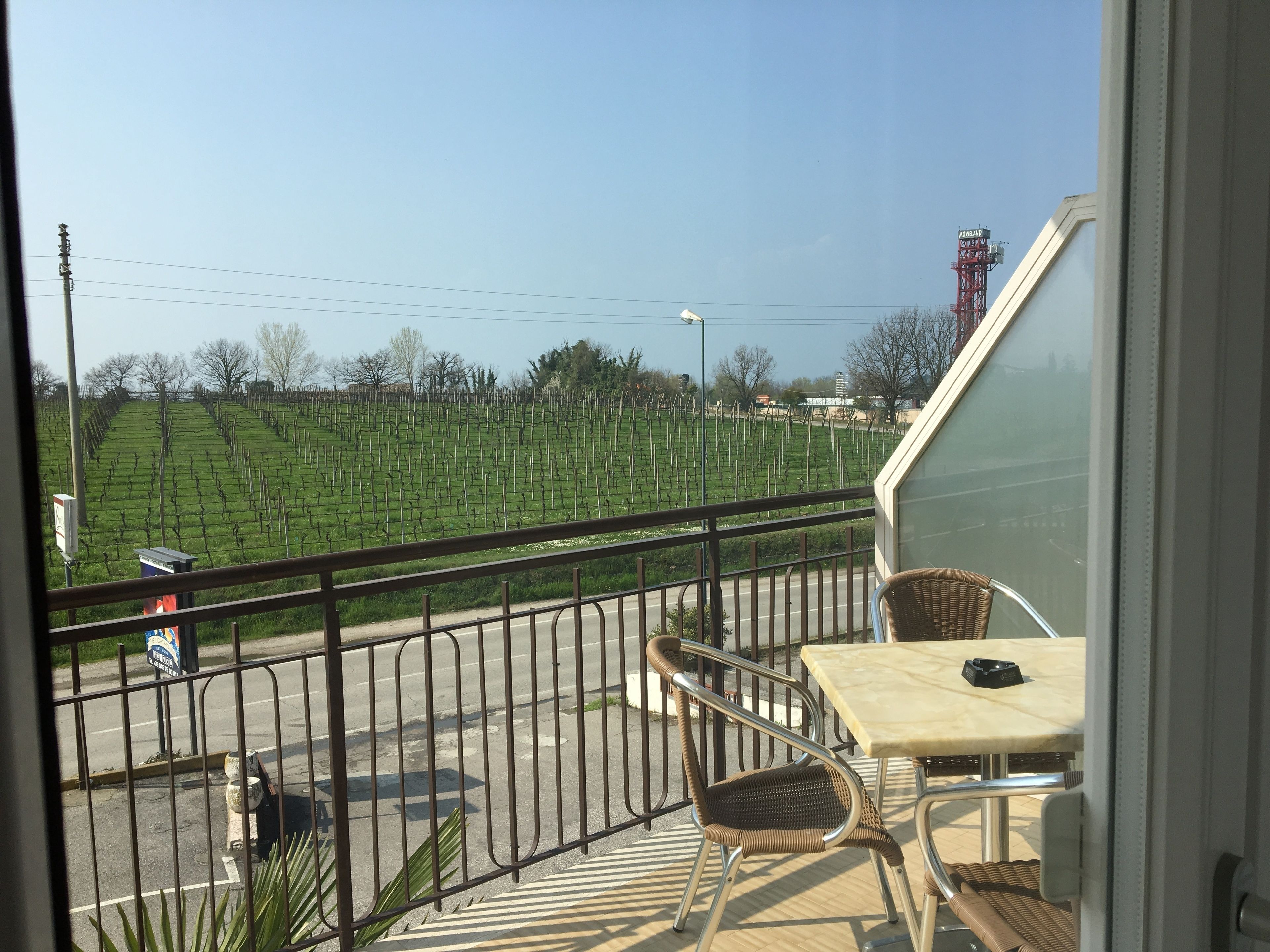 A balcony with a table and two chairs, overlooking a vineyard and a road with a signboard.