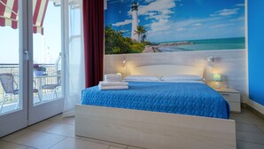 Ein Schlafzimmer mit Bett, Nachttischen, einem Fenster mit Vorhängen und Blick auf einen Strand mit einem Leuchtturm.