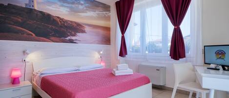 A hotel room with a bed, a desk, a chair, a TV, a nightstand with a lamp, and a large painting of a lighthouse.