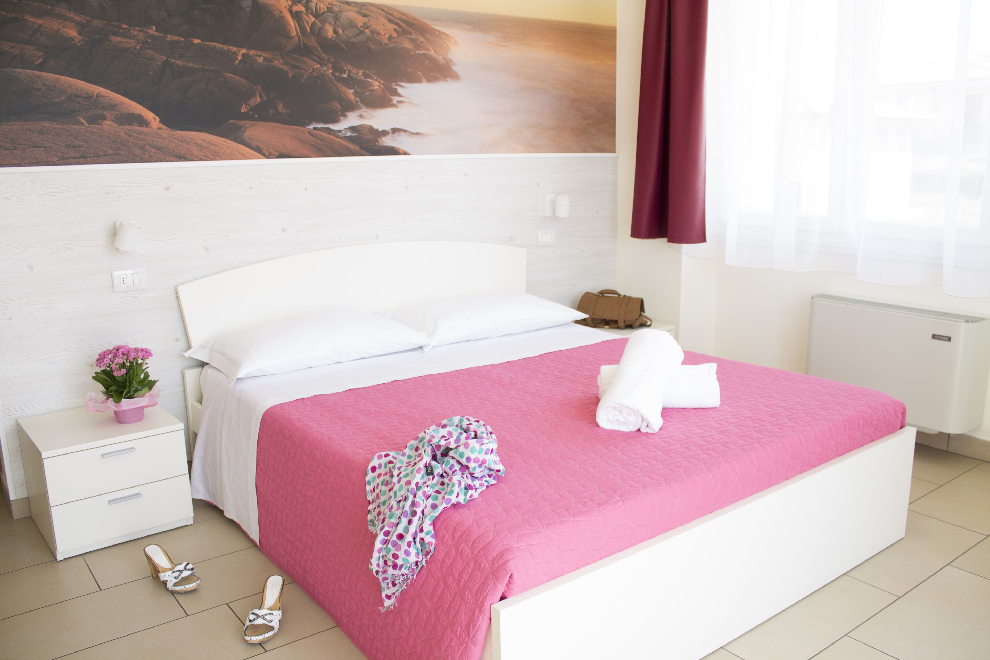 A bedroom with a white bed, pink bedding, a white nightstand, a pink flower vase, and a pair of sandals.