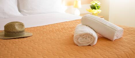 A neatly made bed with white linens, a rolled towel, and a straw hat on an orange quilted bedspread.