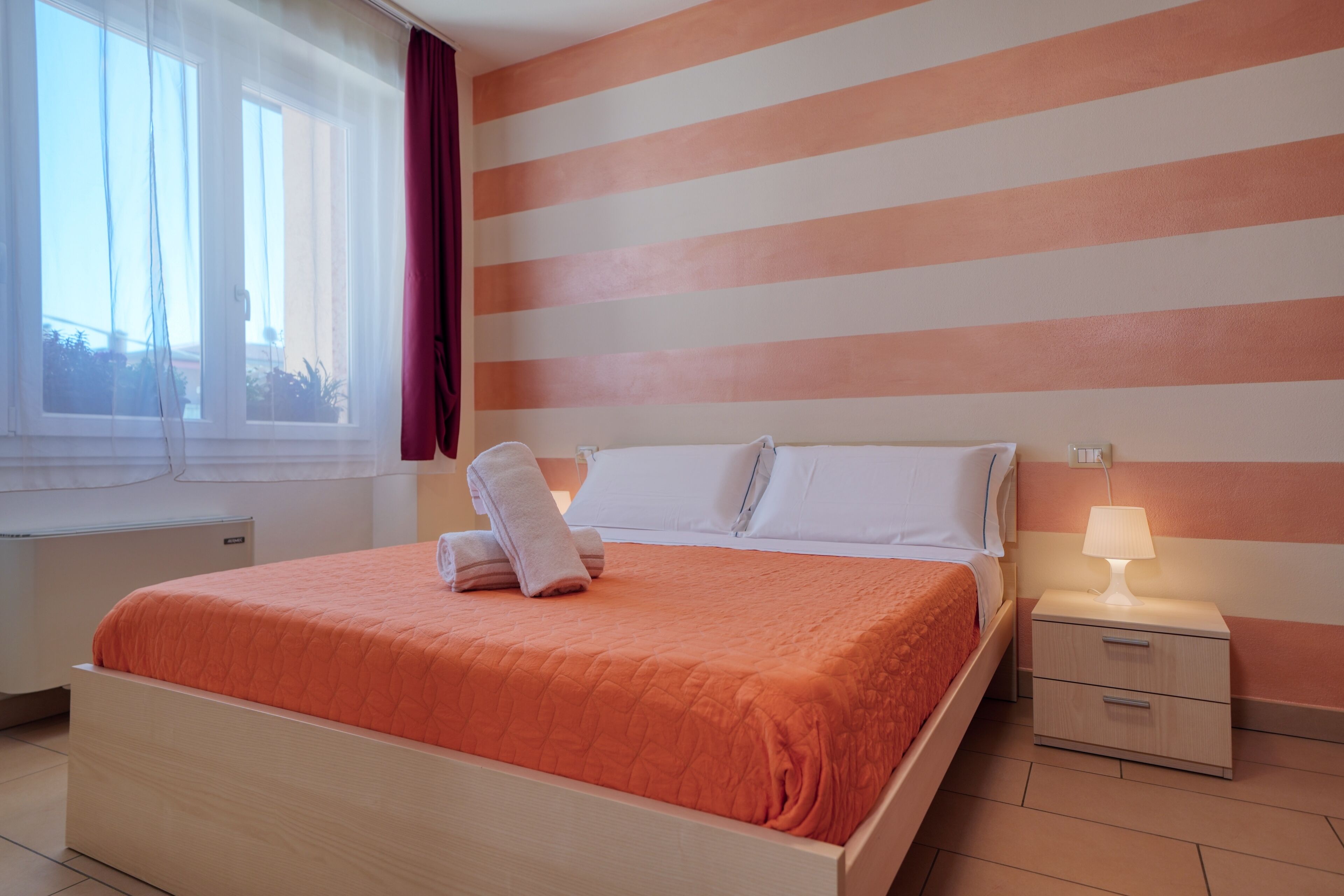 Una cama bien hecha con cabecera blanca y almohadas, una mesita de noche con lámpara, y una ventana con cortinas.