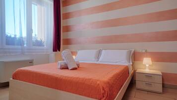 Una cama bien hecha con cabecera blanca y almohadas, una mesita de noche con lámpara, y una ventana con cortinas.