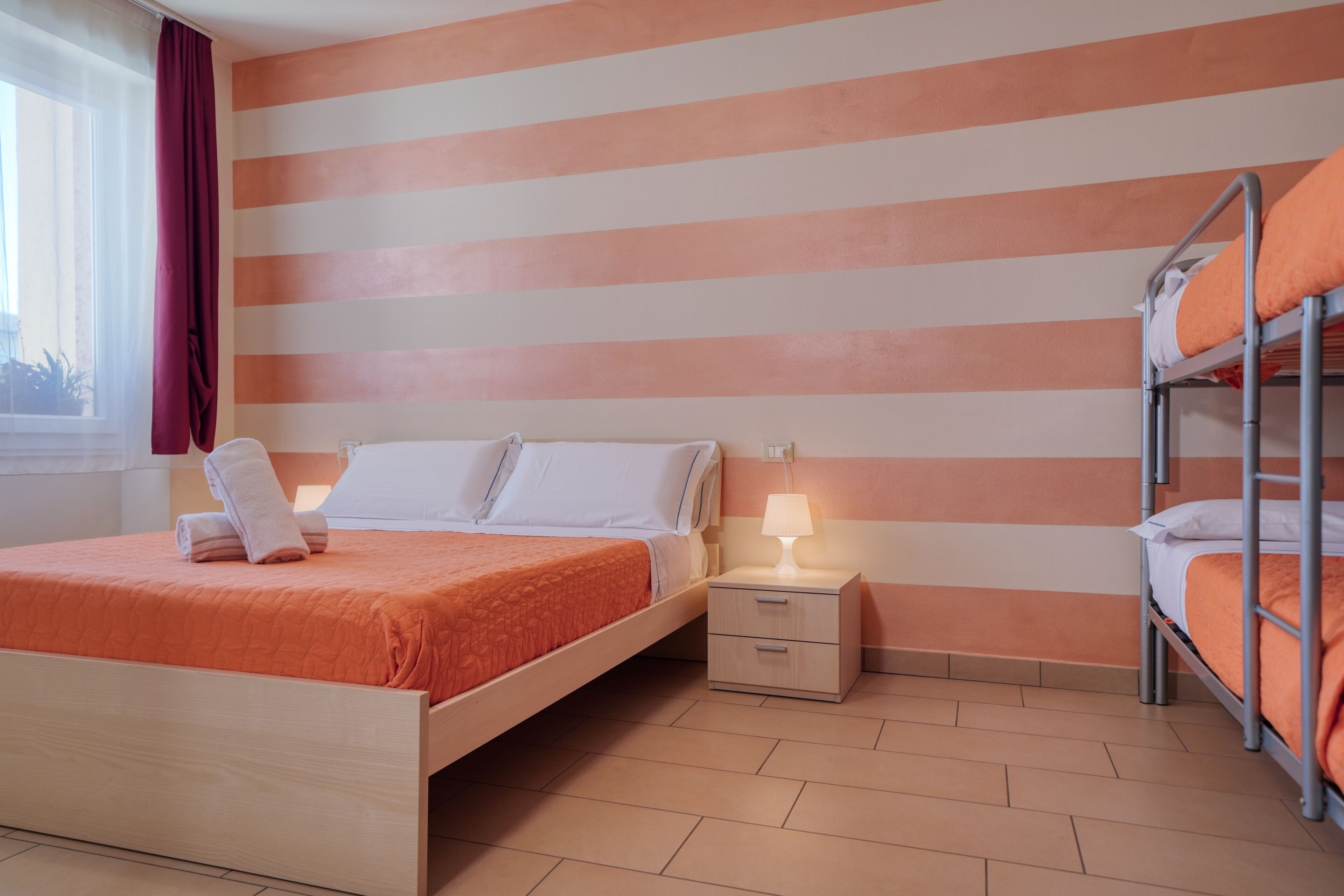 Habitación de hotel con cama de madera, mesita de noche con lámpara, ventana con cortinas y una litera.