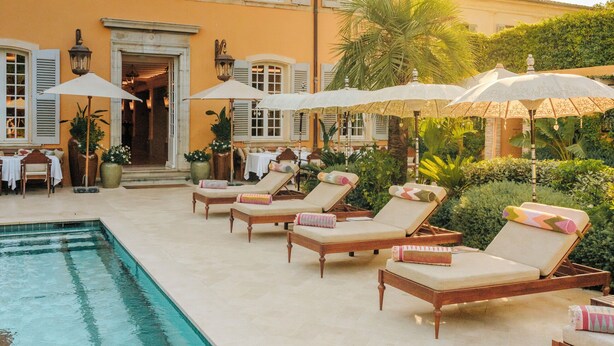 A poolside area with lounge chairs and umbrellas, a building with shutters, and a clear sky. - Airelles Pan Dei Palais (Saint-Tropez)