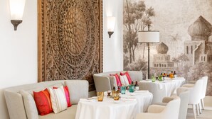A dining area with a long table set for a meal, surrounded by sofas and chairs, with decorative wall panels and a mural in the background. - Airelles Pan Dei Palais (Saint-Tropez)