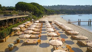 A beachside hotel with rows of sun loungers and umbrellas, a wooden deck, and a view of the sea and hills in the background. - Airelles Pan Dei Palais (Saint-Tropez)