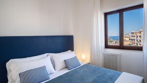 Un lit avec une tête de lit sombre, une literie blanche et des oreillers à rayures. Une fenêtre offre une vue sur des immeubles et un ciel bleu.