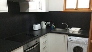 Una cocina compacta con encimera negra, armarios blancos, horno integrado, lavadora y una ventana sobre el fregadero.