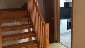 Una escalera de madera que lleva a una puerta oscura, con una cocina visible al fondo.