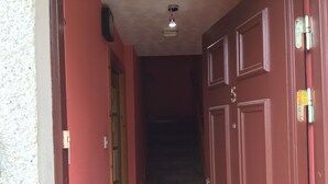 Un couloir avec une porte rouge portant le numéro « 5 » et une porte en bois. On peut voir un luminaire au plafond et une horloge fixée au mur.