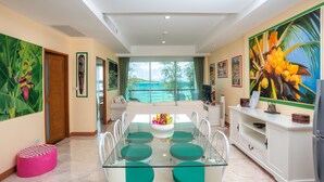 A modern dining area with a glass table, green cushioned chairs, a sofa, and a view of the ocean. - Almali Rawai Beach Residence (Rawai)